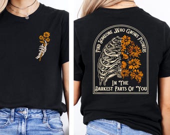 May include: Black t-shirt with a skeleton hand holding yellow flowers on the front. The back of the shirt features a rib cage design with yellow flowers and the text "Find Someone Who Grows Flowers In The Darkest Parts Of You."