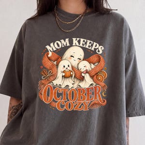 May include: A charcoal gray t-shirt featuring a graphic print of three ghosts wrapped in a large orange scarf. The text reads MOM KEEPS OCTOBER COZY. The design includes illustrations of a mug, cinnamon sticks, and orange slices in warm tones.