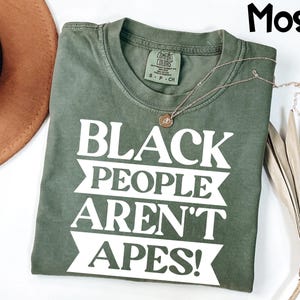 May include: A moss green t-shirt with the text "BLACK PEOPLE AREN'T APES!" in white block letters. A gold necklace with a pendant rests on the shirt. The shirt is folded and displayed with a brown hat and dried plant.