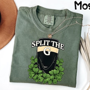 May include: Moss green t-shirt with a graphic of a pint of stout with the text "SPLIT THE G" and shamrocks. The shirt has a crew neck and short sleeves. A gold necklace is draped across the shirt.