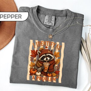 May include: A pepper-colored t-shirt featuring a graphic of a raccoon wearing a beanie and scarf, holding a coffee cup, surrounded by autumn leaves and pumpkins. The text reads I IDENTIFY AS A PROBLEM. The shirt has a crew neck and short sleeves.