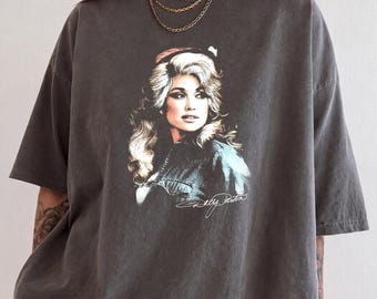 May include: A charcoal gray short-sleeve t-shirt featuring a graphic print of Dolly Parton with blonde hair and a denim jacket. The print includes a white signature script below the portrait. The shirt has a crew neckline and a relaxed fit.