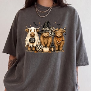 May include: A charcoal gray short-sleeve t-shirt featuring a graphic of three Highland cows dressed for Halloween. One cow wears a ghost sheet with a pumpkin bucket, one wears a witch hat, and one wears an orange bandana, surrounded by bats.