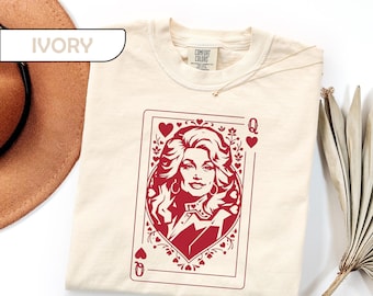 May include: An ivory short-sleeve t-shirt featuring a red graphic print of a playing card with a portrait of a woman with voluminous hair. The design includes heart symbols and the letter Q in the corners. The shirt has a Comfort Colors brand label.