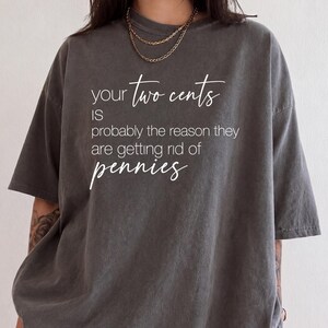 May include: A charcoal grey short-sleeve t-shirt featuring white text that reads: your two cents IS probably the reason they are getting rid of pennies. The shirt has a crew neckline and a relaxed fit, with the text printed in a mix of script and sans-serif fonts.