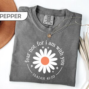 May include: A pepper-colored short-sleeve t-shirt featuring a white daisy graphic with an orange center. The text "fear not, for i am with you" and "ISAIAH 41:10" is printed in white around the flower, accompanied by small four-pointed star icons.