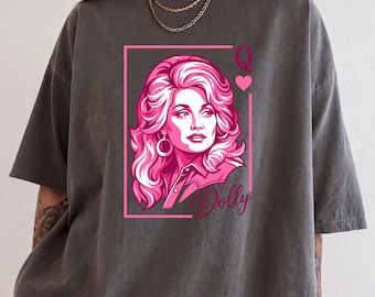 May include: A charcoal gray short-sleeve t-shirt featuring a pink graphic print of a woman with voluminous hair inside a rectangular frame. The design includes the letter Q, a heart symbol, and the cursive text Dolly below the portrait.