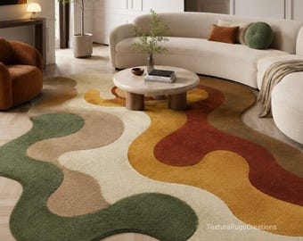 May include: An irregular, organic-shaped area rug featuring a wavy, multi-colored pattern in shades of sage green, cream, mustard yellow, terracotta, and brown. The rug has a textured pile surface and includes the text TexturaRugsCreations.