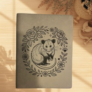 Peut inclure: Un journal gris avec une illustration noire détaillée d'un opossum entouré d'une couronne florale de roses et de marguerites. L'opossum est représenté assis, la queue enroulée. Le dessin est centré sur la couverture du journal.