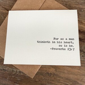 May include: A white greeting card with the text "For as a man thinketh in his heart, so is he. -Proverbs 23:7" in black font. The card is inside a brown envelope, set against a wooden surface.