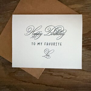 May include: A cream-colored birthday card with the words "Happy Birthday To My Favorite Leo" in elegant black script. The card is placed on top of a brown envelope, set against a wooden surface.