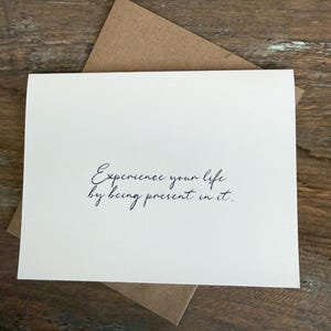 Peut inclure: Une carte blanche avec le texte "Experience your life by being present in it" en écriture noire, posée sur une enveloppe marron. La carte et l'enveloppe sont sur une surface en bois.