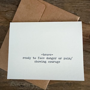 Op de afbeelding: Een witte kaart met het woord "-brave-" en de definitie in het zwart gedrukt. De kaart ligt op een bruine envelop, tegen een houten achtergrond.