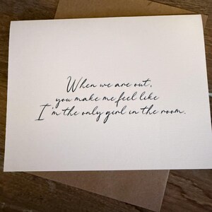 Puede incluir: Una tarjeta de felicitación blanca con el texto manuscrito: "When we are out, you make me feel like I'm the only girl in the room." La tarjeta está sobre un sobre marrón, sobre una superficie de madera.