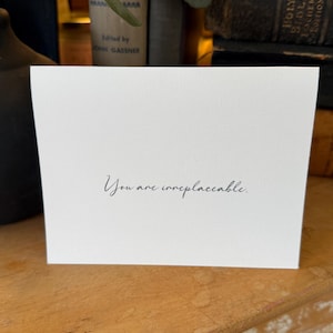 Op de afbeelding: Een witte kaart met de tekst "You are irreplaceable" in sierlijke letters. De kaart staat voor een verzameling oude boeken, wat een vintage esthetiek creëert, perfect voor een cadeau of een speciale boodschap.