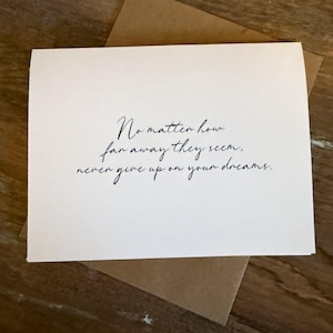 May include: A white greeting card with the handwritten text "No matter how far away they seem, never give up on your dreams." The card is on a brown envelope, with a wooden background.