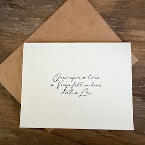 May include: A cream-colored card with the text "Once upon a time a Virgo fell in love with a Leo" in black script, inside a brown envelope. The card and envelope are on a wooden surface.