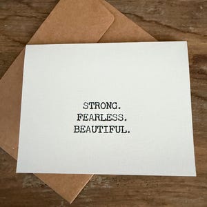 Puede incluir: Una tarjeta blanca con las palabras "STRONG. FEARLESS. BEAUTIFUL." impresas en negro, colocada sobre un sobre marrón. La tarjeta y el sobre están sobre una superficie de madera.