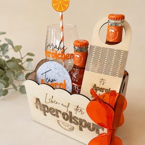 Könnte beinhalten: Eine Holzkiste mit einem Glas mit der Aufschrift Sarahs Sprölchen, zwei Aperol-Flaschen, einem grauen Filzuntersetzer mit der Aufschrift Team Aperol, einem hölzernen Flaschenträger mit der Aufschrift it's Aperol time, einem Orangenscheiben-Strohhalm und orangefarbenen herzförmigen Brillen.