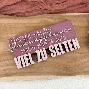 Könnte beinhalten: Ein rechteckiges Schild in Mauve mit dem deutschen Text "EINFACH MAL INS GLÜCKSNÄPFCHEN MACHT MAN JA AUCH VIEL ZU SELTEN" in weißer und rosa Schrift. Das Schild liegt auf einem Holztablett, mit rosa Stoff und Blumen im Hintergrund.