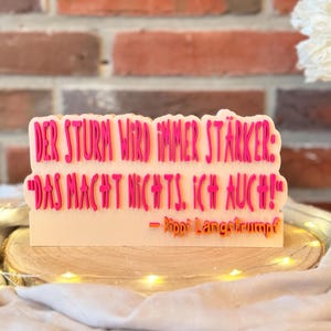 Könnte beinhalten: Dekoratives Schild mit dem Zitat "DER STURM WIRD IMMER STÄRKER: 'DAS MACHT NICHTS. ICH AUCH!' - Pippi Langstrumpf" in rosa Schrift. Das Schild steht vor einem Ziegelhintergrund und auf einem Holzsockel.