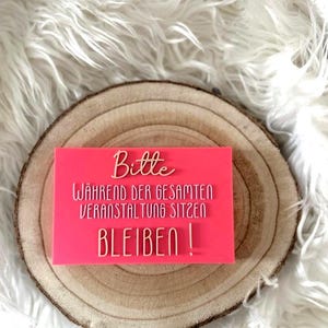 Op de afbeelding: Een felroze rechthoekig bord met gouden letters met de tekst "Bitte, Während der gesamten Veranstaltung sitzen, BLEIBEN!", geplaatst op een houten schijf. De achtergrond is een zacht, wit oppervlak.