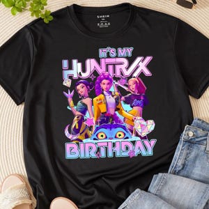 May include: Black crew neck t-shirt with a colorful graphic. The graphic features three animated women and the text "IT'S MY HUNTRAX BIRTHDAY" in a gradient font. The shirt is made of a soft material.