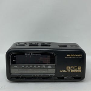 May include: A black Soundesign AM/FM/Weather Band clock radio. The radio has a digital display with FM and AM frequency indicators. The text "Instant Weather" is visible on the front, along with control buttons for volume, power, and band selection.