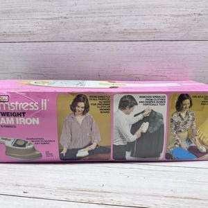 May include: A pink box for a "Steamstress II" lightweight steam iron. The box features images of the iron in use and text that reads "Irons directly on all fabrics" and "Removes wrinkles from clothes and drapes."