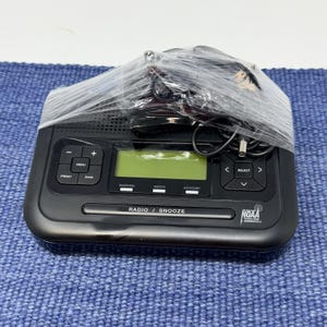 May include: A black digital radio alarm clock with a green LCD screen, wrapped in clear plastic. The clock has buttons for menu, preset, band, and select. The text "RADIO / SNOOZE" is visible.