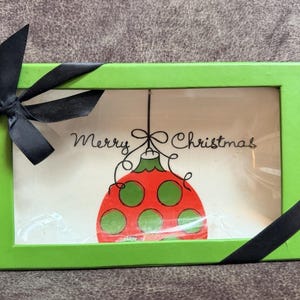 May include: A rectangular green box with a black bow and ribbon. The box contains a card with a red and green Christmas ornament design and the words "Merry Christmas" in script.