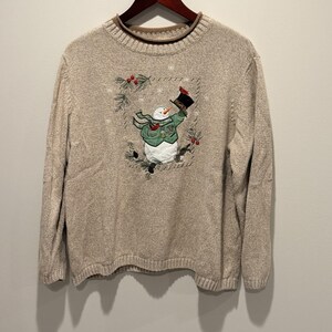 May include: Beige long-sleeve sweater with a snowman design. The snowman wears a black top hat, green jacket, and a scarf. The design is framed by pine branches and red berries. The sweater has a ribbed hem and cuffs.