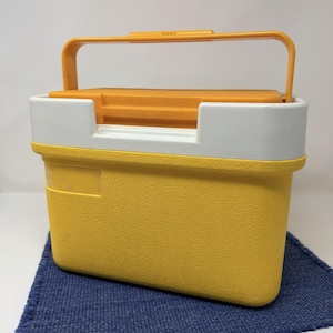 May include: A yellow and white plastic cooler with an orange handle and lid. The cooler has a textured surface and a rectangular shape. The handle is arched and attached to the top of the cooler. The cooler is sitting on a blue cloth.