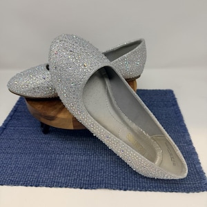 May include: A pair of silver-colored, rhinestone-covered ballet flats. The shoes are displayed on a small wooden stand atop a blue woven mat. The flats have a rounded toe and a low heel.