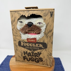 Könnte beinhalten: Ein brauner Pappkarton mit einem ausgeschnittenen Gesicht eines Monsters mit weißem Fell, rosa Lippen und einem braunen Schnurrbart. Auf der Box steht "ZURU Fuggler Funny Ugly Monster" und "Hairy Fuggs".