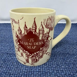 Puede incluir: Taza de cerámica color crema con una ilustración granate de un castillo y las palabras "The Marauder's Map". La taza tiene un asa curva y la palabra "Hogwarts" es visible en una pancarta. La taza está sobre una superficie azul texturizada.