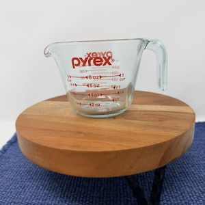 Puede incluir: Un vaso medidor Pyrex transparente con marcas rojas para onzas y tazas. El vaso tiene un asa y está sobre un pequeño soporte redondo de madera con patas de metal negro. El soporte está sobre una superficie azul.