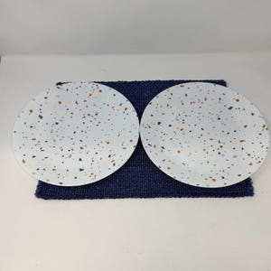May include: Two white ceramic plates with a speckled design in blue, brown, and grey. The plates are round and rest on a dark blue woven placemat. The plates are a modern addition to any kitchen.