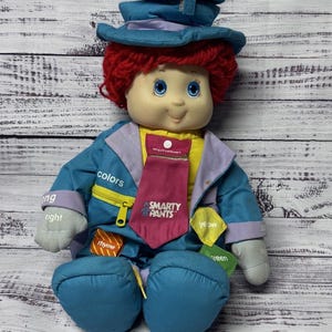 以下が含まれることがあります： 赤い毛糸の髪、青い目、笑顔の表情をした、ヴィンテージのSmarty Pants人形。人形は青いジャケット、黄色のシャツ、ピンクのネクタイを着ています。ジャケットには「colors」と「rhyme」という言葉が書かれています。