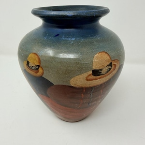 May include: A hand-painted ceramic vase with a blue rim and a rounded body. The vase features a design of two figures wearing hats, painted in shades of brown, red, and blue. The vase is a decorative item.