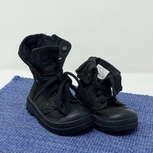 May include: Pair of black canvas boots with laces and a rubber sole. The boots have a high-top design with a folded-down collar. The boots are sitting on a blue surface.