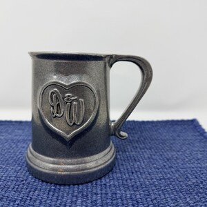 May include: A pewter-colored mug with a heart-shaped emblem featuring the intertwined letters "D&W." The mug has a sturdy handle and a slightly flared base. The background is a blue textured surface.
