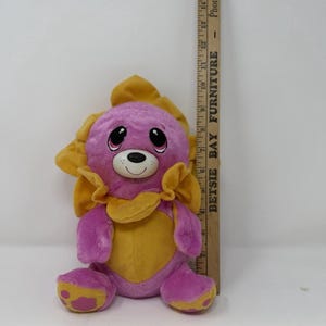 May include: A plush toy in the shape of a lion with a pink body and a yellow mane and chest. The toy has a smiling face with black eyes and a white nose. The toy is approximately 8 inches tall.