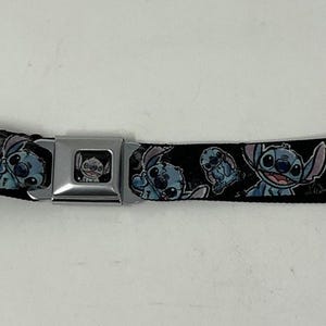 May include: A black seatbelt featuring a repeating pattern of the character Stitch from the animated film Lilo & Stitch. The belt has a silver metal buckle and is laid flat against a white surface. The Stitch characters are in various poses.