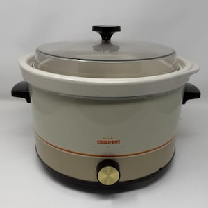 Op de afbeelding: Crème-kleurige Rival Crock-Pot slowcooker met een doorzichtig glazen deksel en zwarte handgreep. De basis heeft een bruine band met een oranje streep en een goudkleurige knop. De tekst "RIVAL CROCK-POT" is zichtbaar.