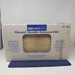 Puede incluir: Una caja rectangular de filtro de repuesto para humidificador Bemis Waterwick. La caja es blanquecina con texto azul y un filtro beige visible a través de una abertura rectangular. El texto incluye "Wicking filter for clean evaporative humidifying traps mineral deposits."