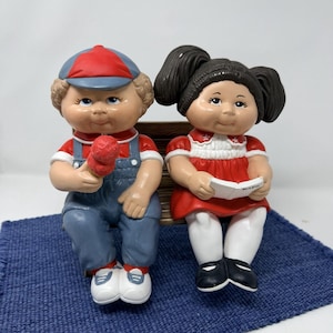 May include: A pair of vintage ceramic figurines of children sitting on a bench. The boy wears a red and blue cap, red shirt, blue overalls, and holds a red popsicle. The girl wears a red dress and holds a book. The bench sits on a blue mat.