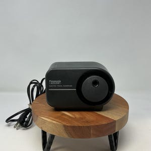 May include: A black Panasonic electric pencil sharpener with the text "AUTO-STOP" and "ELECTRIC PENCIL SHARPENER". The sharpener is on a small, round wooden stand with black metal legs. The power cord is visible.