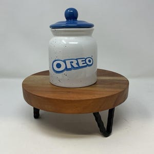 Op de afbeelding: Een witte keramische Oreo-koekjespot met een blauw deksel en het Oreo-logo in blauw. De pot is gespikkeld en staat op een ronde houten standaard met zwarte metalen poten.
