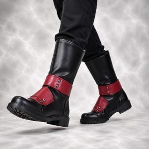 May include: A pair of mid-calf boots in black with red ankle straps. Each boot features a rounded toe, a thick black sole with a treaded bottom, and two silver-toned metal studs on each red strap. The boots are worn with black trousers.